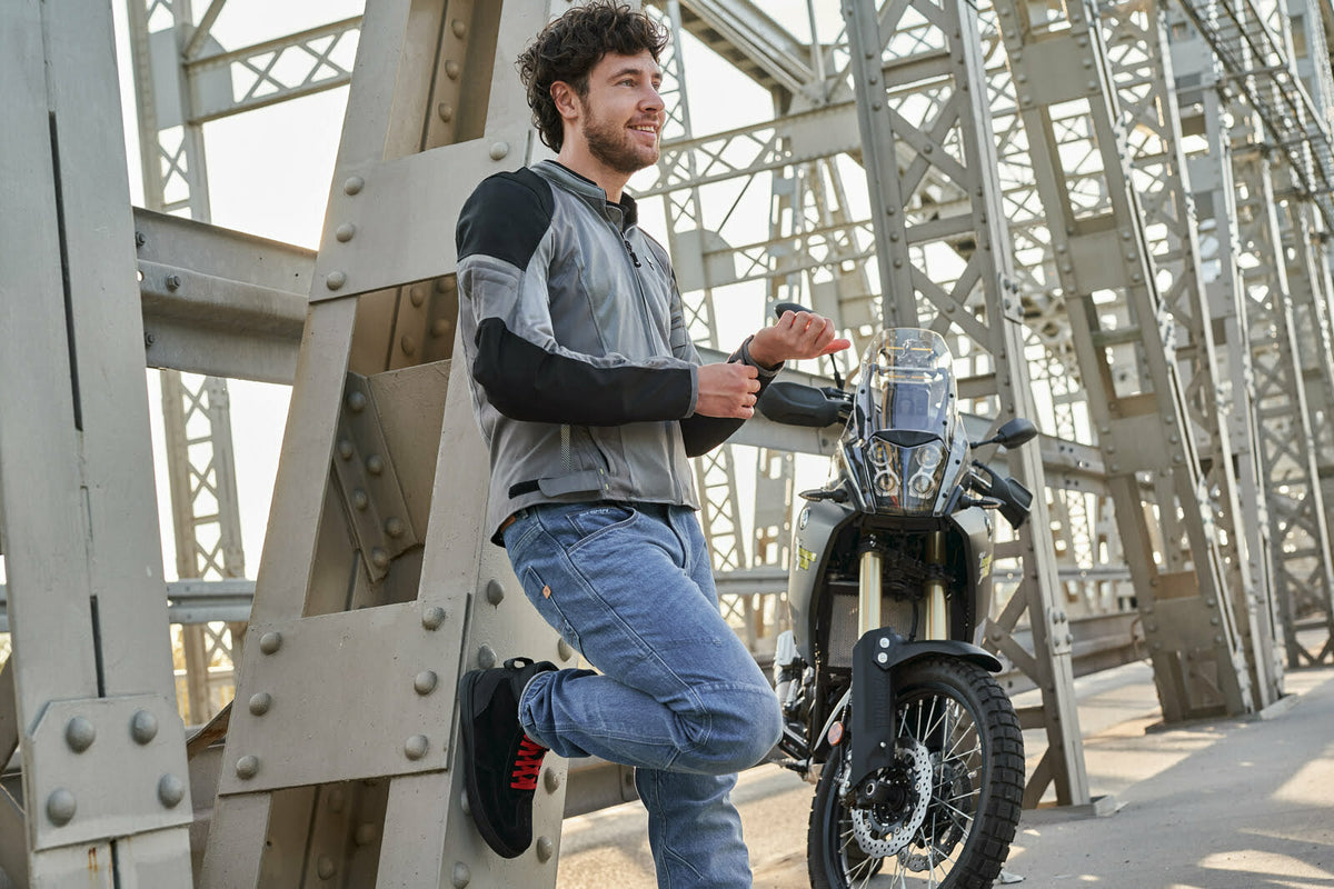 Man bär Shima Kevlar Mc-Jeans Devon Blå vid motorcykel på bro. Kombination av skydd och stil för motorcykelentusiaster.